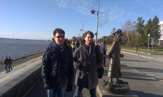 Два молодых человека стоят около карикатруного памятника Чехову на набережной в Томске